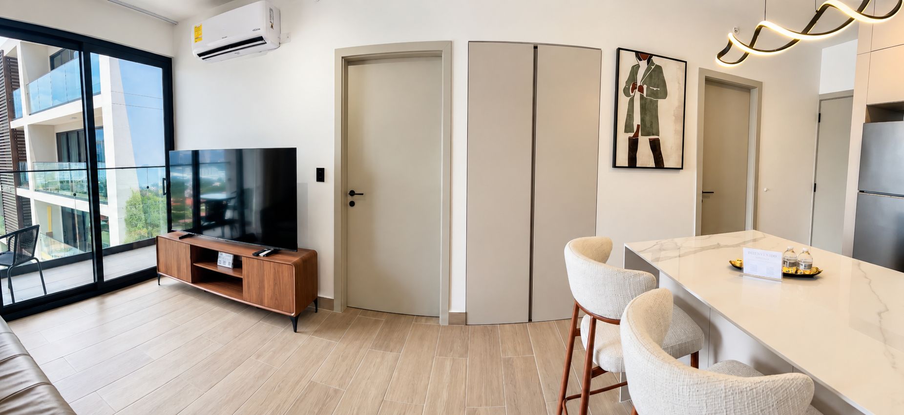 A modern living room with a flat-screen TV, a wooden entertainment unit, a dining area with bar stools, and a large window offering a view of the outdoors.