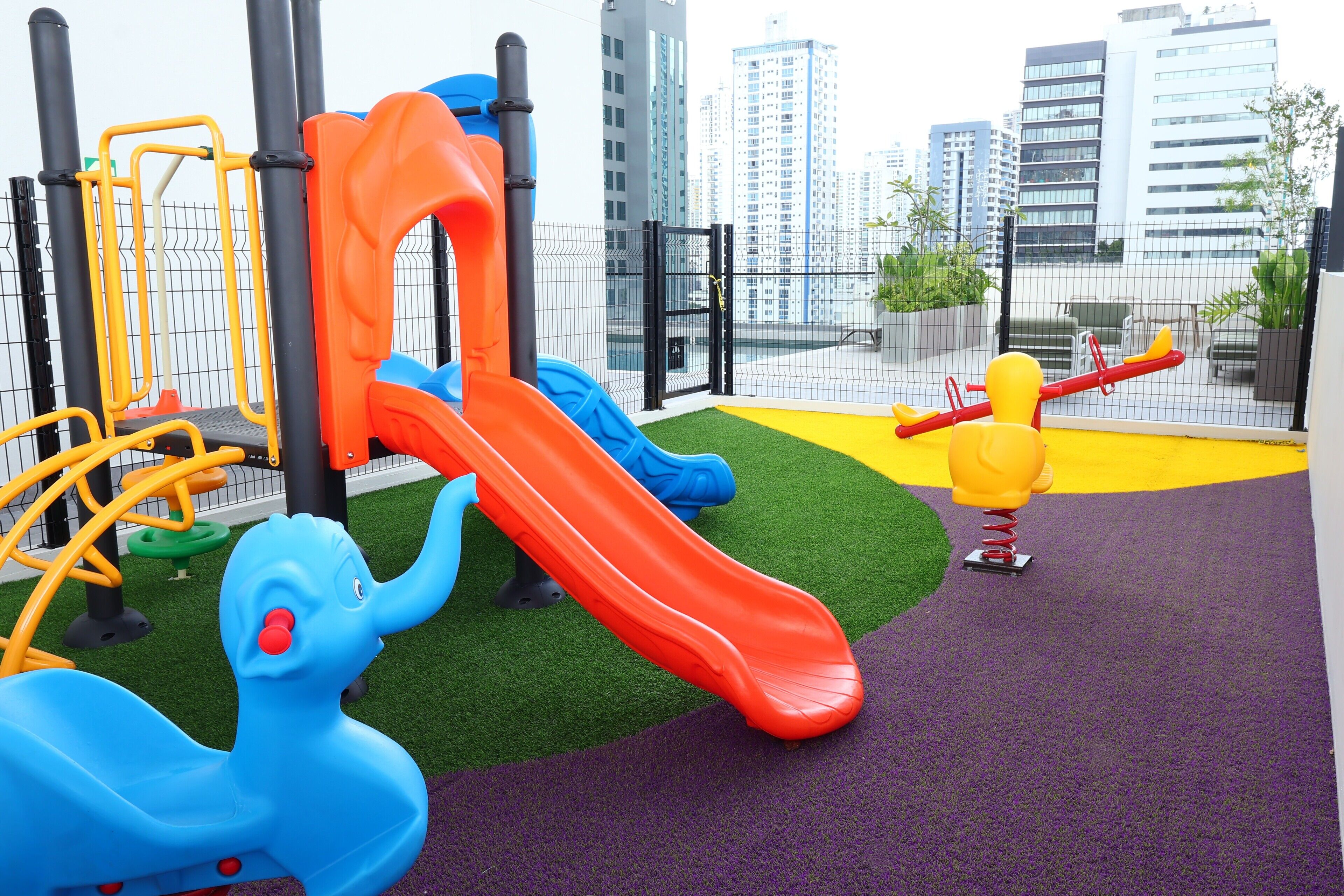 A playground with slides and climbing structures, surrounded by a fenced area with a view of tall buildings.