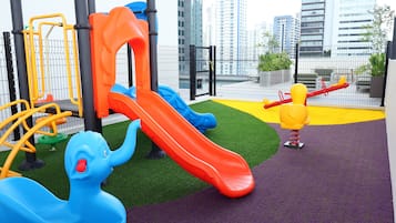 A playground with slides and climbing structures, surrounded by a fenced area with a view of tall buildings.