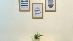 Ein kleiner Essbereich mit einem Holztisch, zwei weißen Hocker, einer Topfpflanze und drei gerahmten Bildern an der Wand.