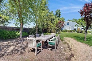 Une table en bois, entourée de chaises, se trouve dans un coin du jardin, avec une grange en arrière-plan.