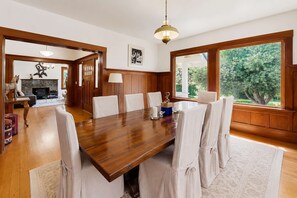 A dining room with a wooden table, white chairs, a chandelier, and large windows with a view of greenery.