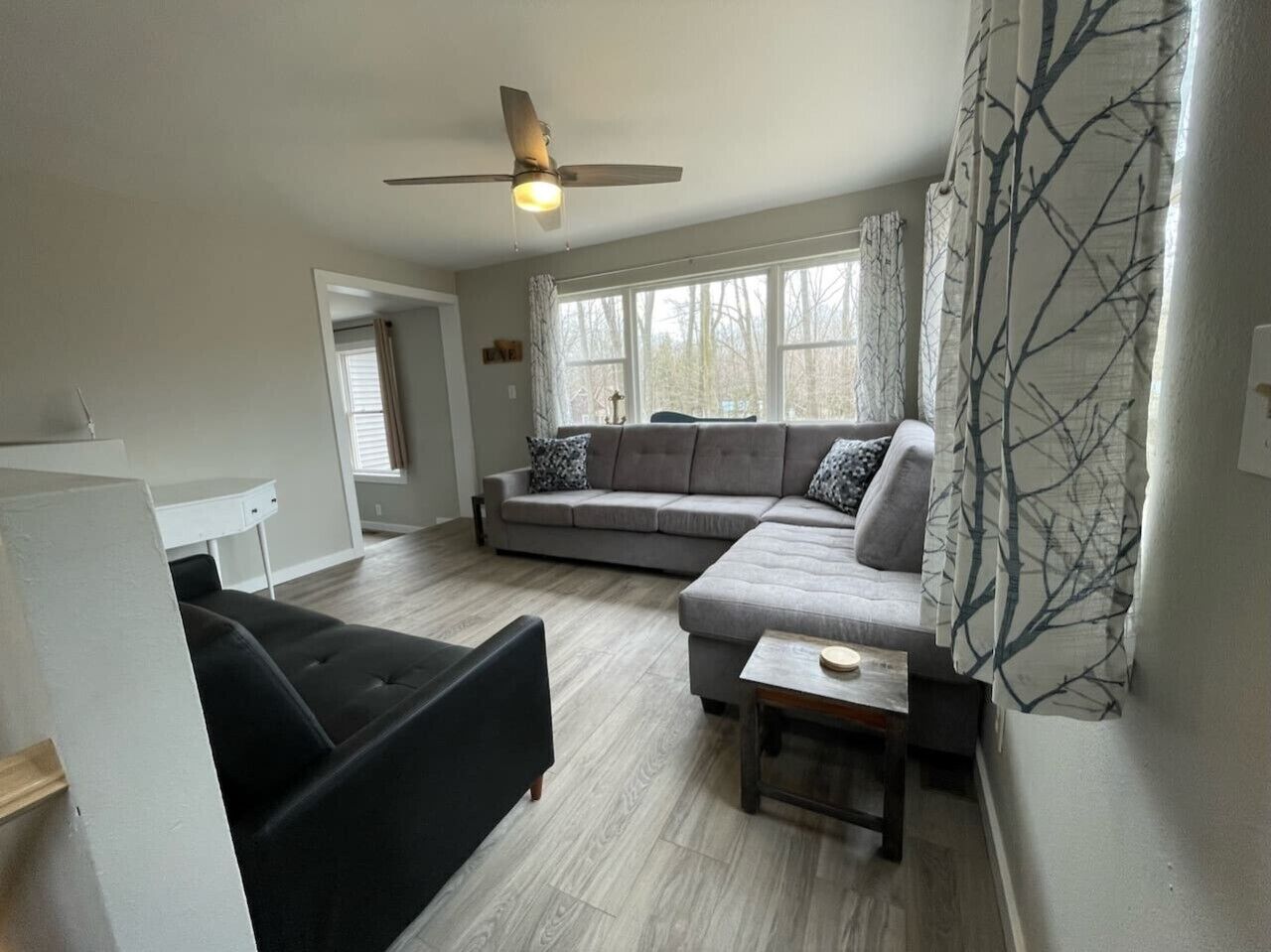 A living room with a sofa, a black chair, a wooden coffee table, a ceiling fan, and large windows.
