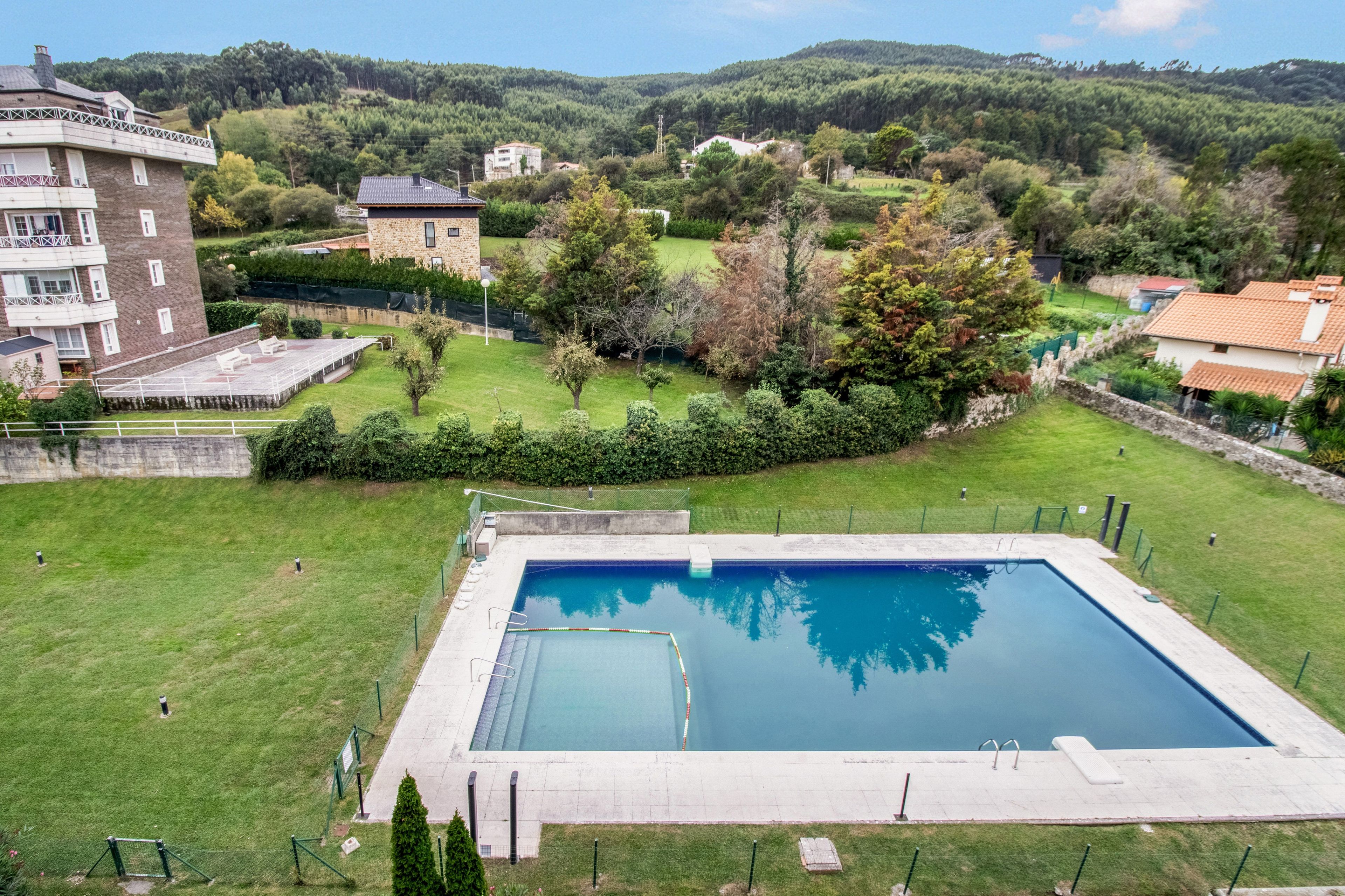 Una piscina rodeada por un césped cuidado, con vistas a edificios residenciales y una colina boscosa al fondo.