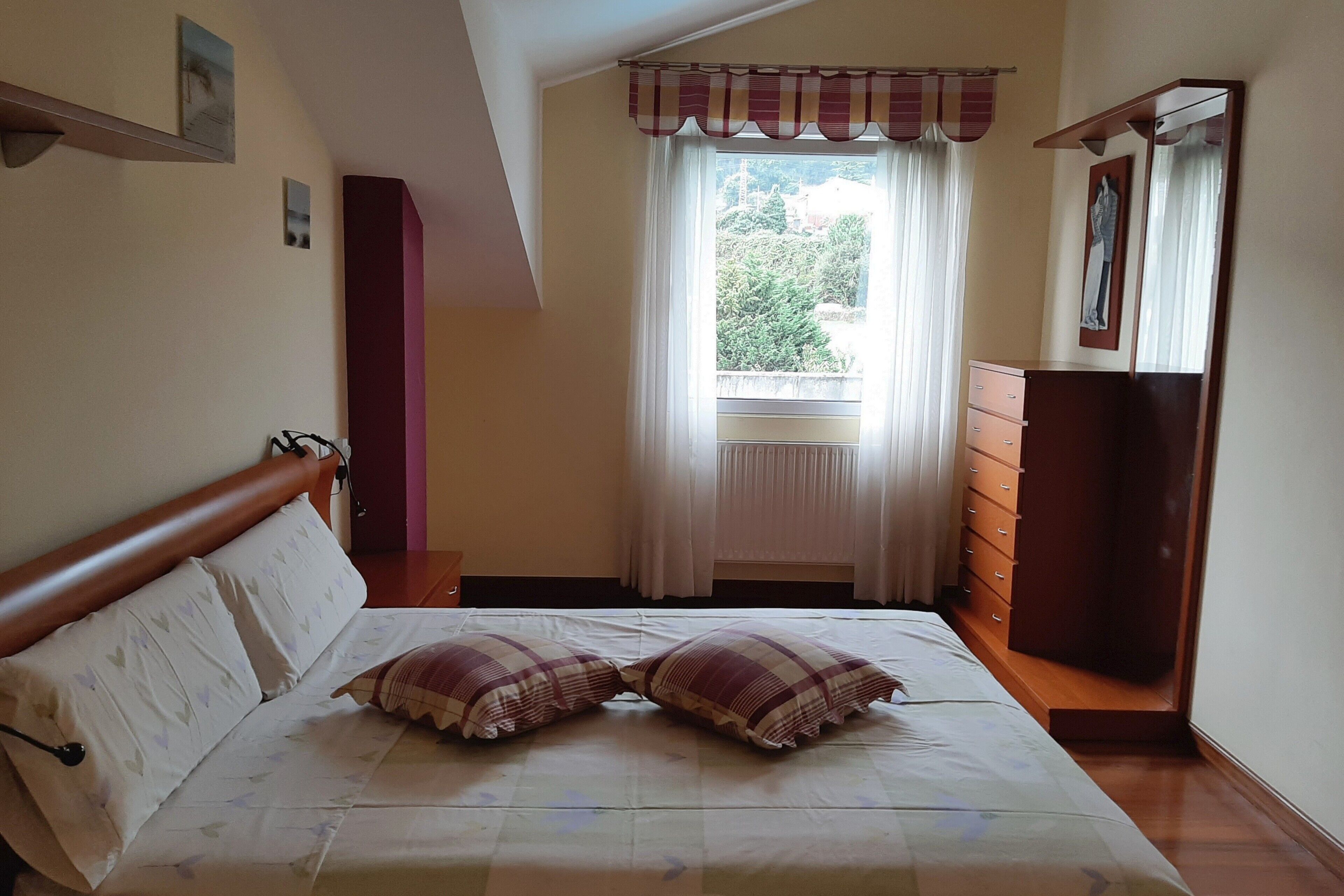 Ein Schlafzimmer mit Bett, Schminktisch, einem Fenster mit Vorhängen und Blick auf Grünfläche.