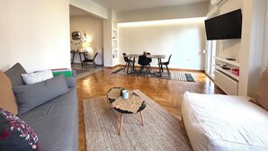Uma sala de estar moderna com um sofá, uma mesa de centro, uma área de refeições com cadeiras e uma TV fixada na parede.