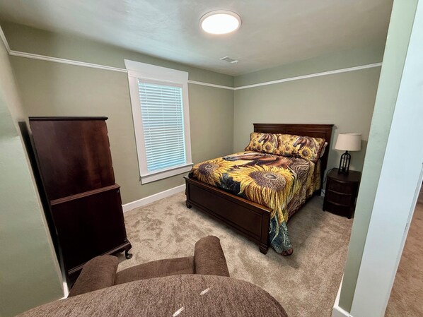 Une chambre à coucher comprenant un lit, une commode, une table de chevet, une lampe et une fenêtre avec des stores.