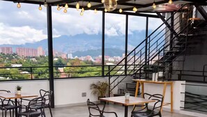 An open-plan dining area with tables and chairs, a staircase, and a view of buildings and mountains. - Comuna 13 Urbana | 1-minute walk to the metro + Coworking (Medellín)