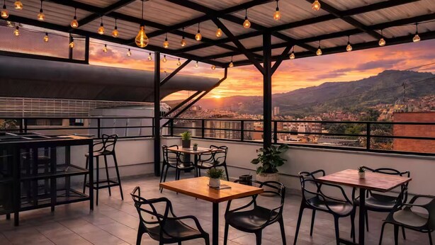 A rooftop terrace with tables and chairs, a view of a sunset over a cityscape, and a metal roof structure. - Comuna 13 Urbana | 1-minute walk to the metro + Coworking (Medellín)