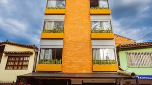 A multi-story building with a central orange brick section, balconies with plants, and a storefront with outdoor seating. - Comuna 13 Urbana | 1-minute walk to the metro + Coworking (Medellín)