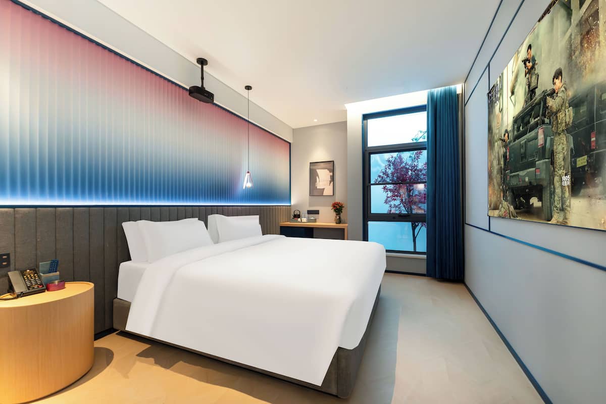 Moderni hotellihuone, jossa on suuri sänky, yöpöytä, jossa on puhelin, pieni työpöytä, ikkuna, josta on näkymä, ja seinä, joka on koristeltu seinämaalauksella.