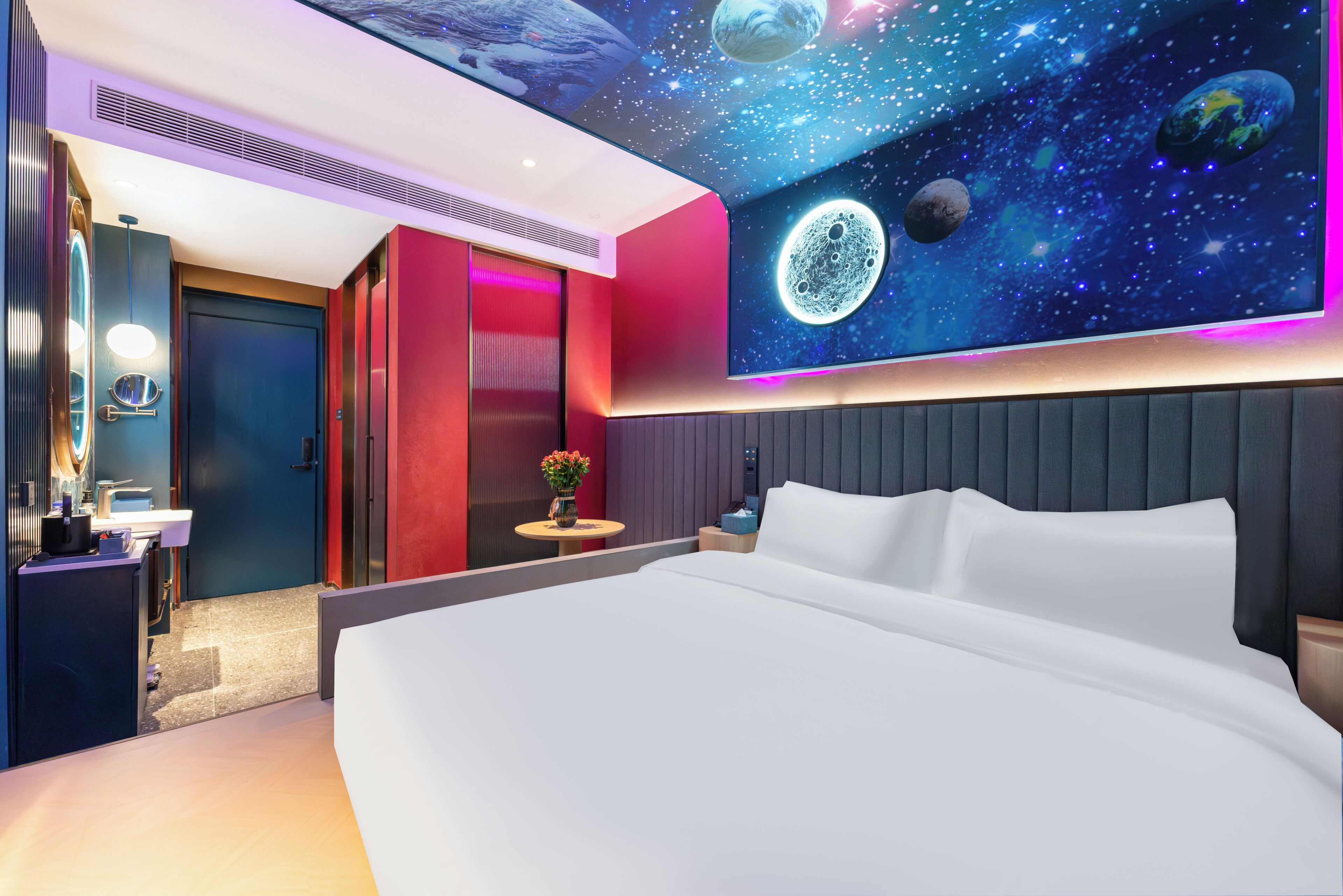 A modern hotel room with a large bed, a vanity with a mirror, a small table with a vase, and a wall mural of space.