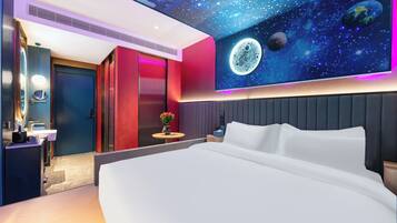 A modern hotel room with a large bed, a vanity with a mirror, a small table with a vase, and a wall mural of space.