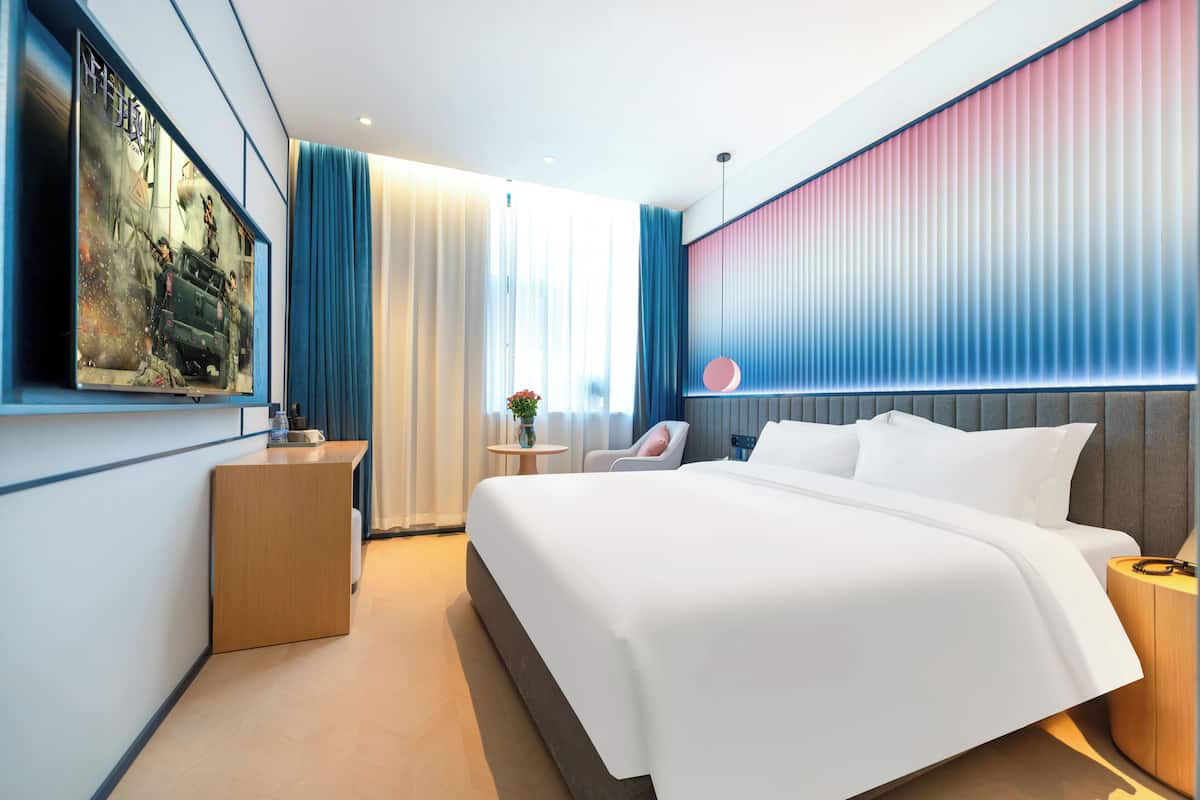 Moderni hotellihuone, jossa on suuri sänky, työpöytä, tuoli, pieni pöytä, jossa on kasvi, ja seinälle asennettu televisio.