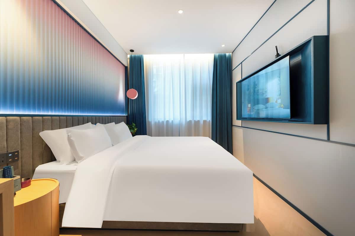 Moderni hotellihuone, jossa on suuri sänky, seinälle asennettu taulutelevisio ja yöpöytä, jossa on lamppu.