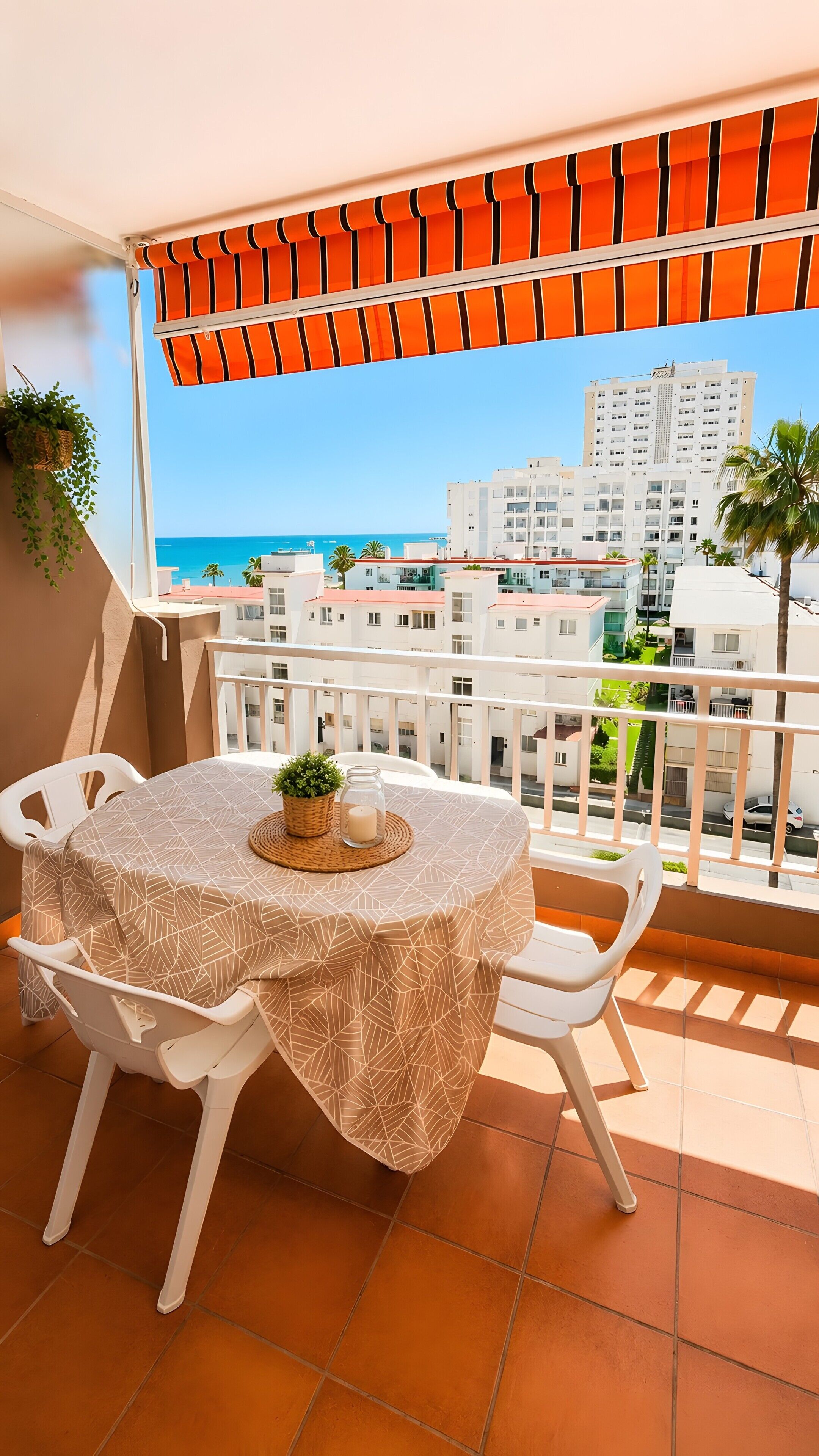 Un balcón con mesa y sillas, vista al mar y edificios al fondo.