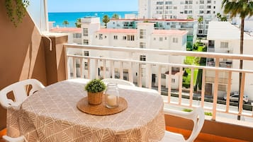 Un balcón con mesa y sillas, vista al mar y edificios al fondo.