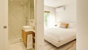 Une salle de bain moderne avec une douche à parois de verre, un meuble-lavabo en bois et un lavabo blanc. La chambre comprend un grand lit avec une literie blanche, une fenêtre avec des rideaux et un appareil de climatisation fixé au mur.