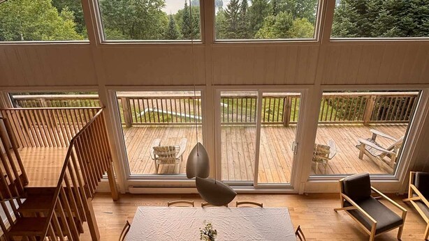 Ruime kamer met een grote tafel en stoelen, een houten trap en ramen met uitzicht op een terras en bergen.