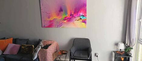 A living room with a sofa, armchair, and a colorful abstract painting on the wall.