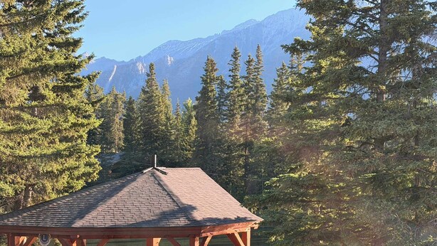 A wooden gazebo with a tiled roof overlooking a lake, surrounded by tall trees and mountains in the background.