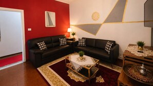 Ein Wohnzimmer mit roter Akzentwand, einem schwarzen Ecksofa mit gemusterten Kissen, einem runden Couchtisch mit weißem Tischläufer und einem geometrischen Wandmuster.