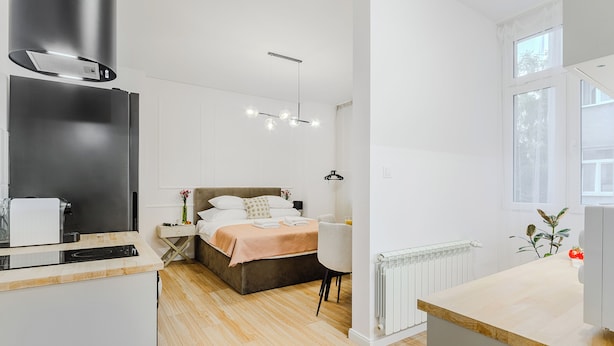 Ett modernt hotellrum med ett litet kök, en säng med sänggavel, ett sängbord, en stol och ett fönster med utsikt över träd.