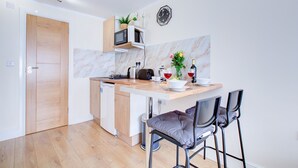 A compact kitchen with a wooden door, a built-in microwave, a small sink, and a marble backsplash. There are two bar stools and a small table with a vase of flowers and wine glasses. - Cozy Twin bed Studio- North London- Free Parking (Enfield)