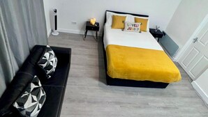 Quarto com cama, mesa de cabeceira, luminária, puf e sofá preto com almofada estampada.