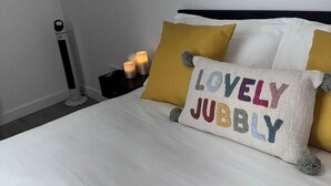 Cama bem arrumada com um travesseiro com a etiqueta 'LOVELY JUBLY' e uma mesa de cabeceira com velas.
