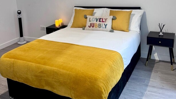 Uma cama com um cobertor amarelo, duas almofadas com texto, uma mesa de cabeceira com uma luminária e uma pequena mesa com uma vela.