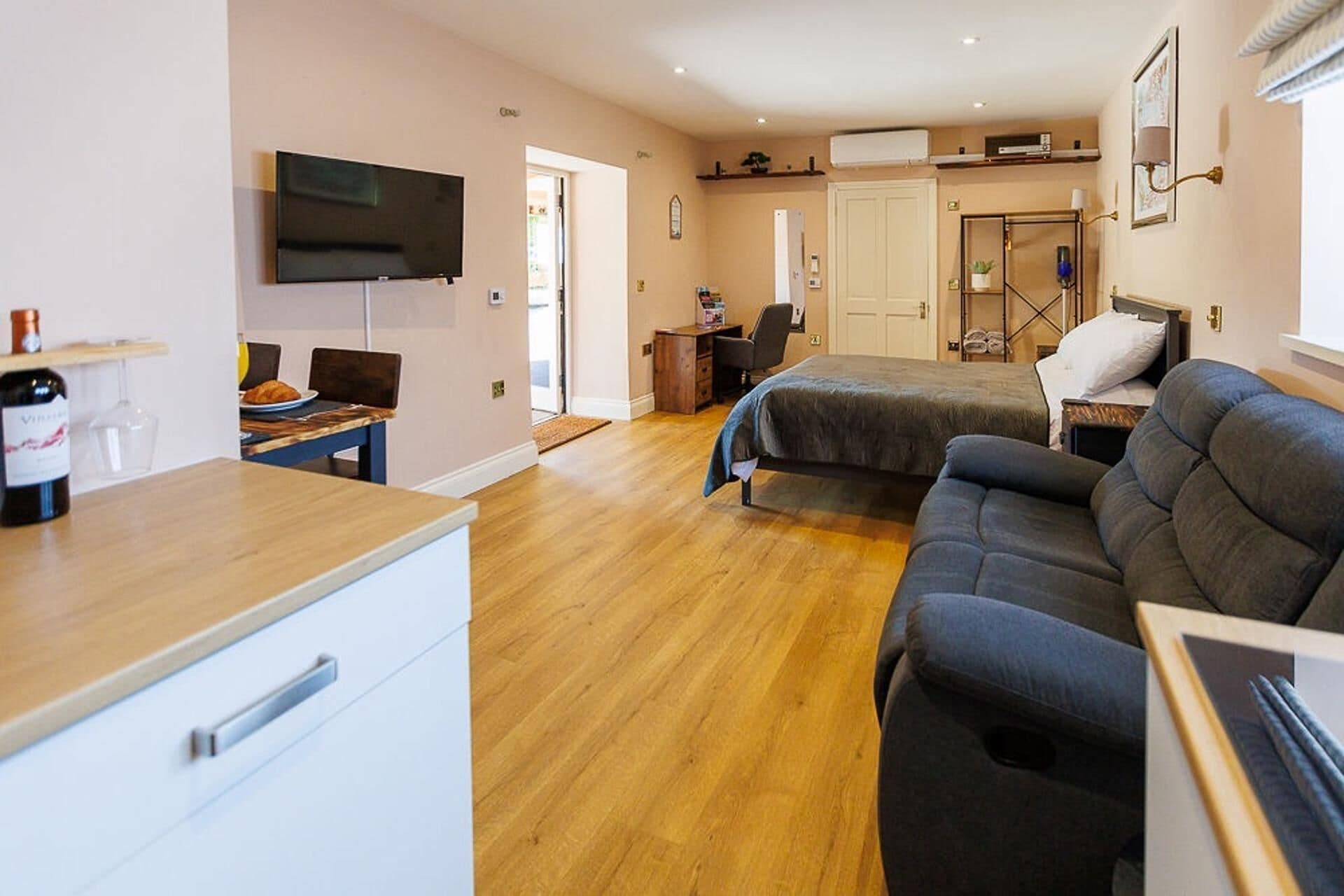 A room with a bed, a sofa, a desk with a chair, a TV mounted on the wall, and a door leading to another room.