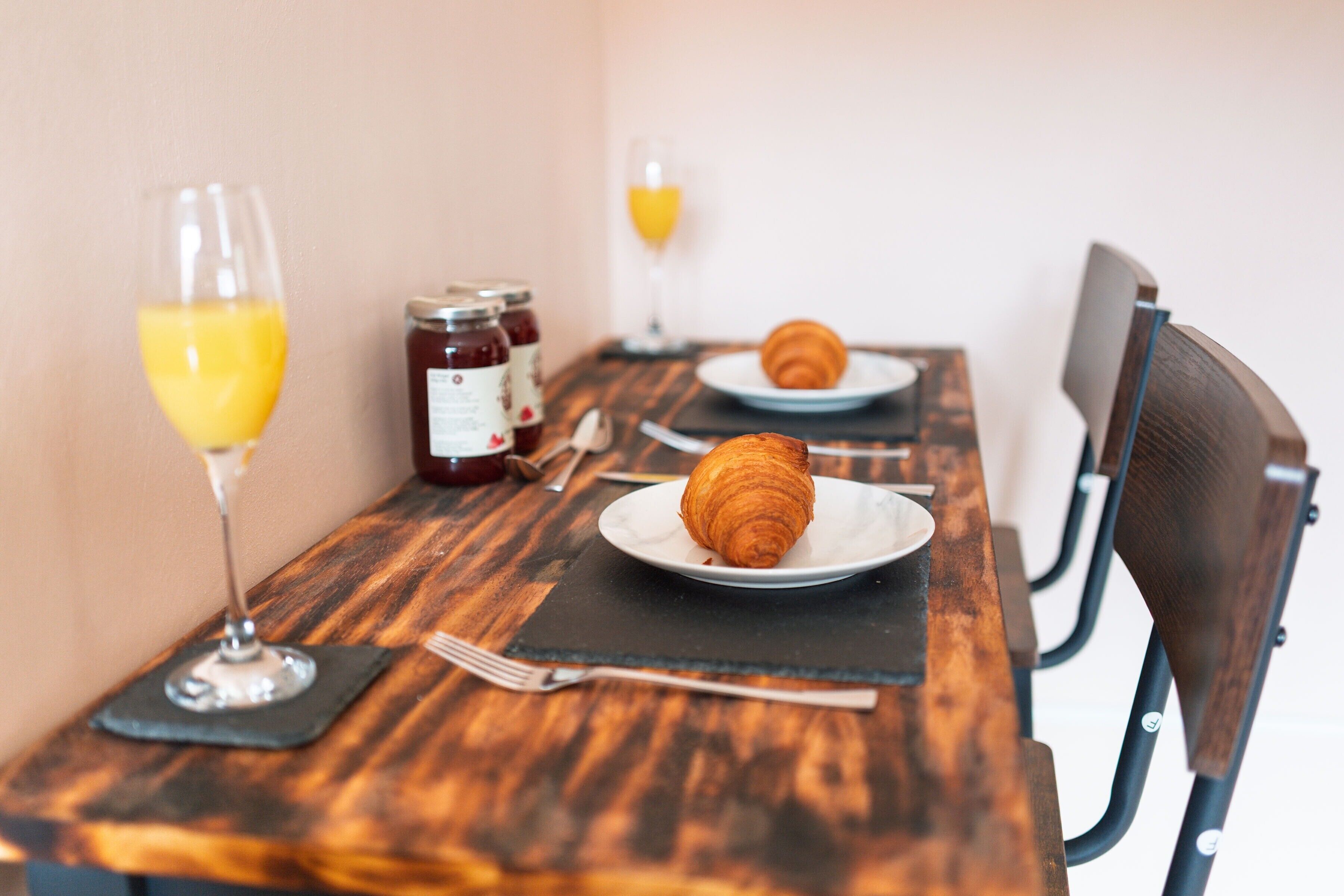 A wooden table with two plates of croissants, a glass of orange juice, and a jar of jam.