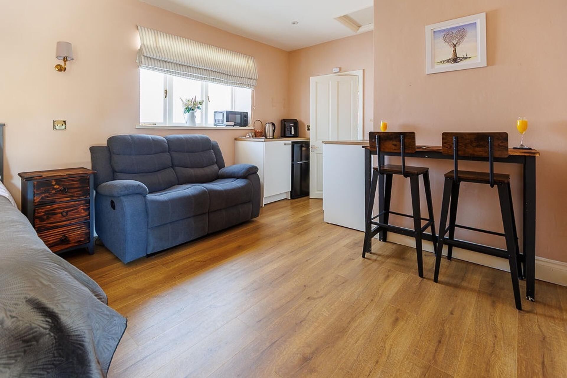 A compact living space with a sofa, a dining area with stools, a kitchenette, and a window with blinds.