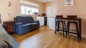 A compact living space with a sofa, a dining area with stools, a kitchenette, and a window with blinds.