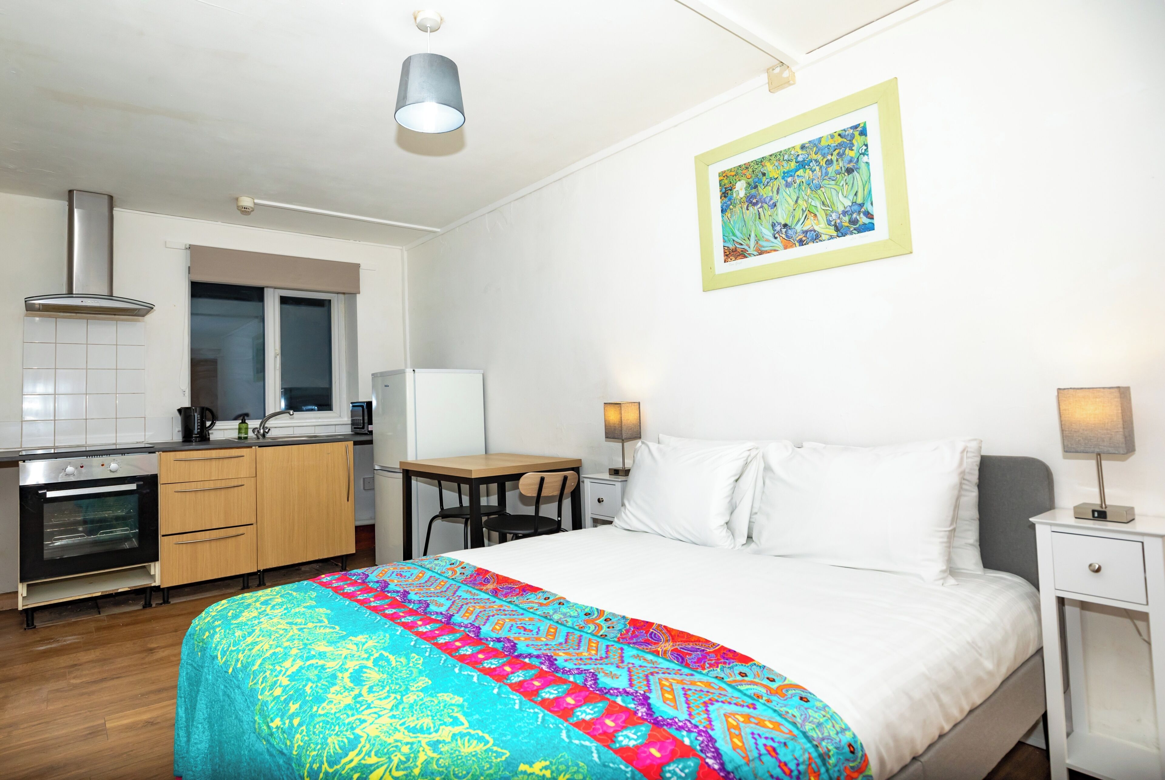 Une chambre d’hôtel compacte comprenant un lit, une table de chevet, un bureau, une chaise, une kitchenette et un tableau au mur.
