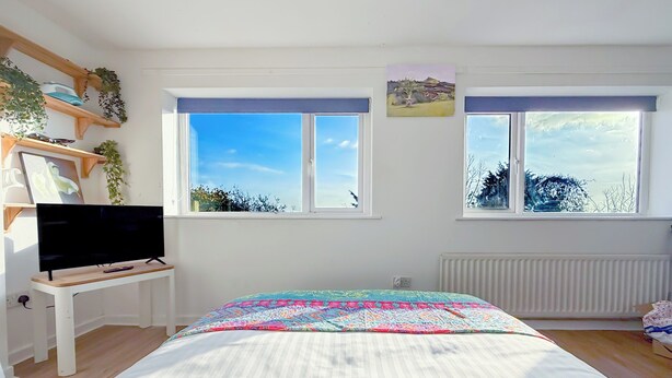 A bedroom with a bed, a TV on a stand, two windows, and a radiator. - Glastonbury Studio- Amazing Views and Free Parking (Glastonbury)