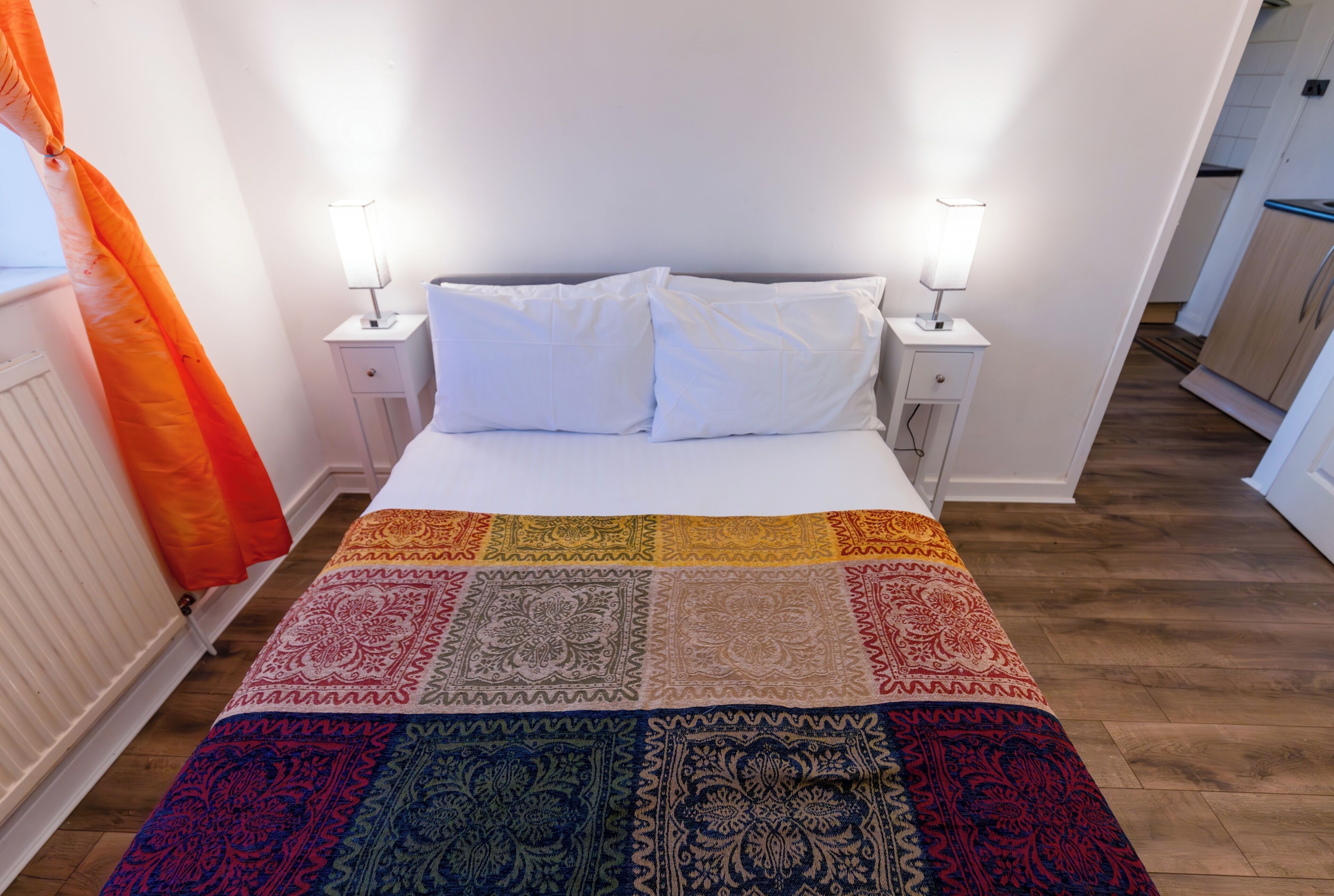 A bedroom with a bed, white pillows, two bedside lamps, a colorful quilt, and a window with an orange curtain.