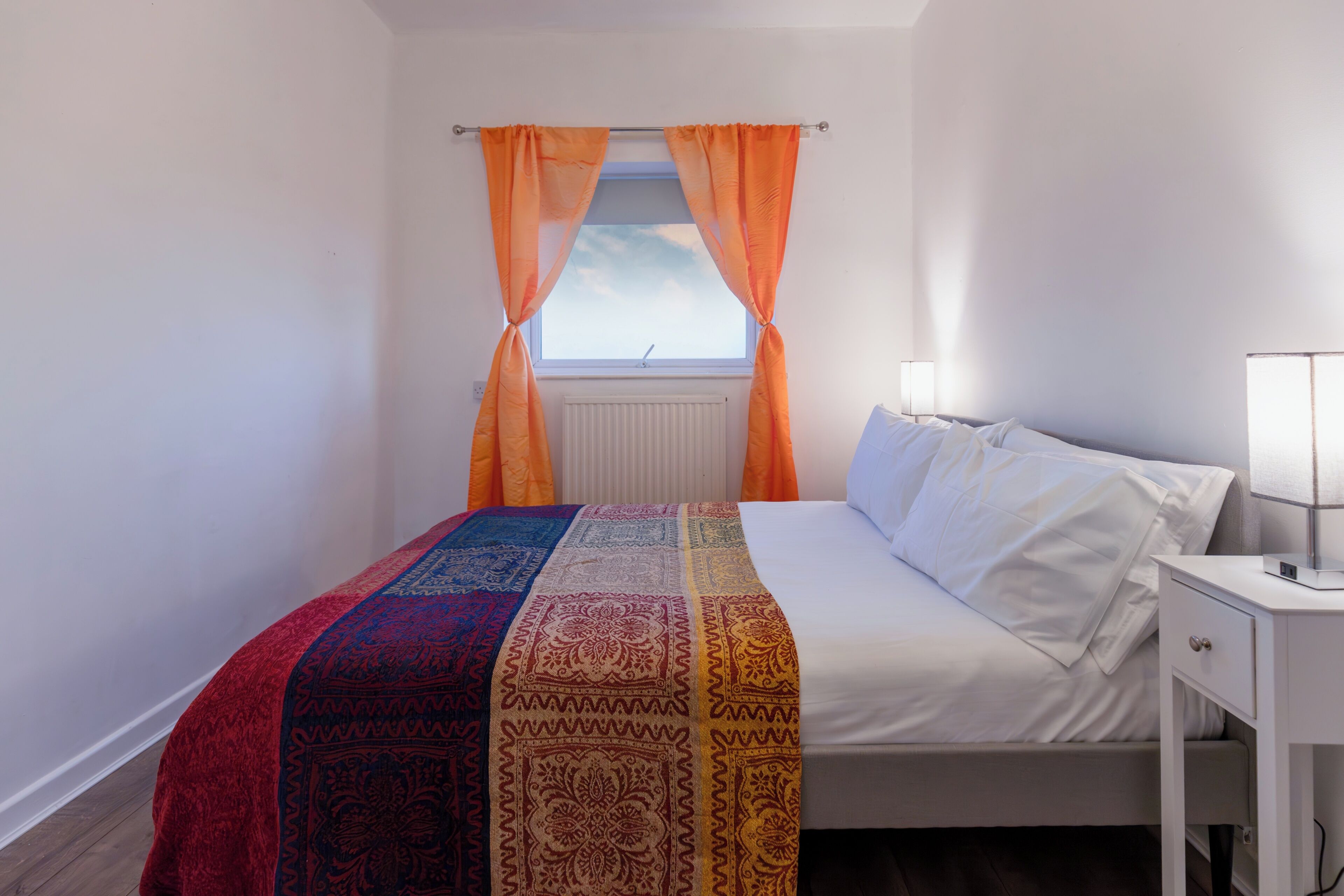 A bedroom with a bed, white curtains, a window, a bedside table with a lamp, and a patterned bedspread.