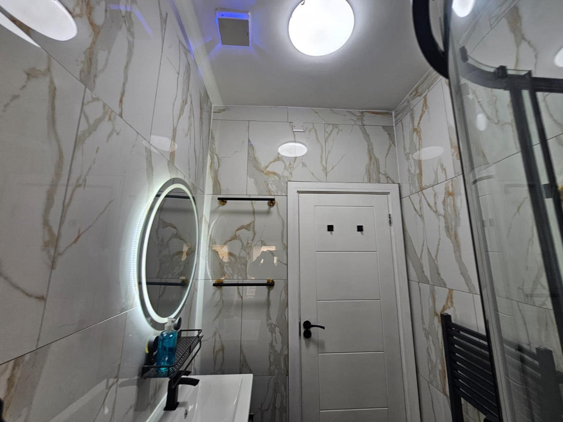 Une salle de bain aux murs recouverts de marbre, une porte blanche, un miroir avec un éclairage intégré et une étagère contenant des articles de toilette.