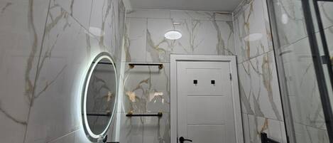 Une salle de bain aux murs recouverts de marbre, une porte blanche, un miroir avec un éclairage intégré et une étagère contenant des articles de toilette.