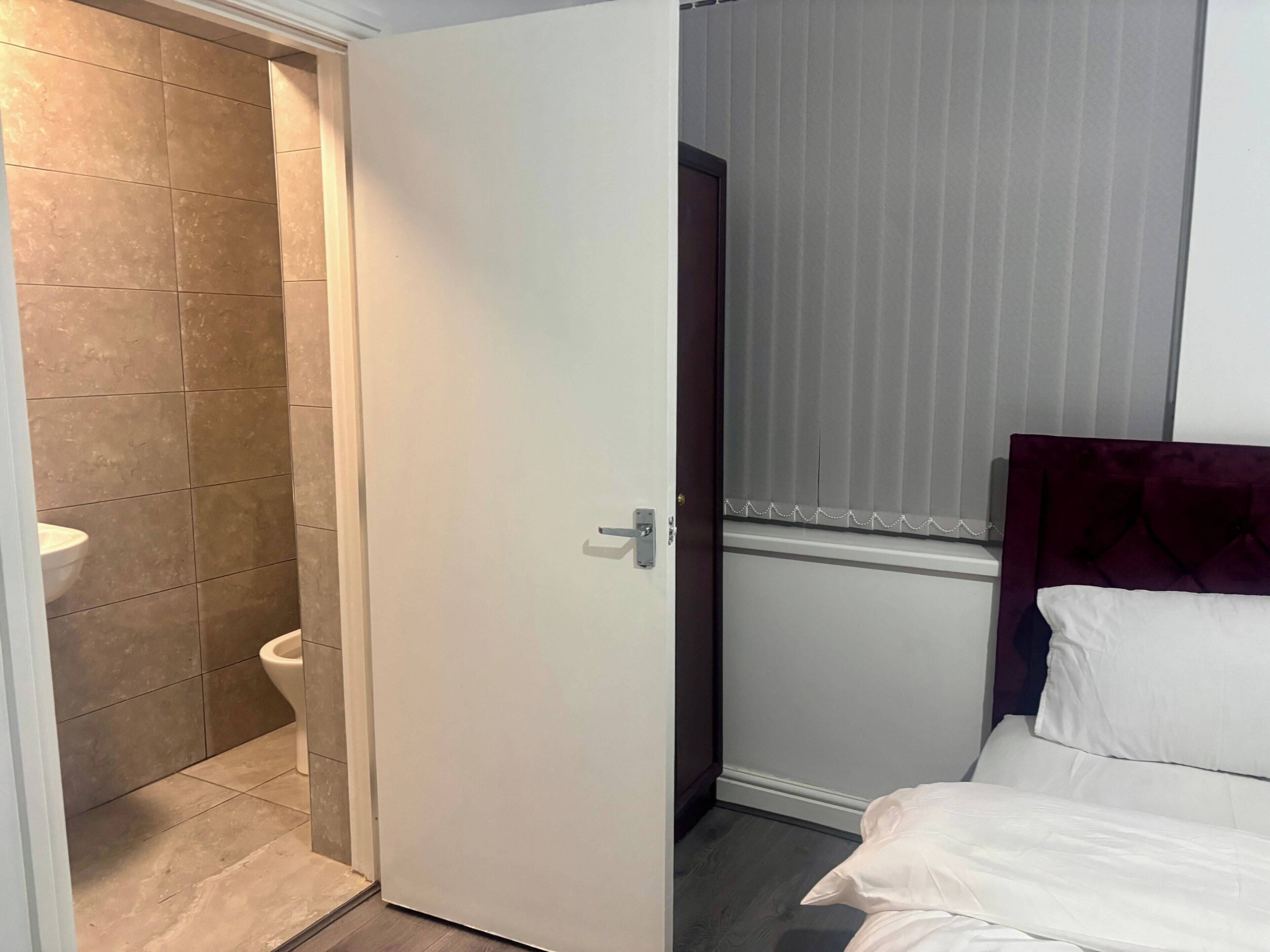 Hotellihuone, jossa on wc, valkoisilla lakanoilla varustettu sänky ja ikkuna, jossa on sälekaihtimet.