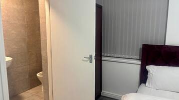 Hotellihuone, jossa on wc, valkoisilla lakanoilla varustettu sänky ja ikkuna, jossa on sälekaihtimet.