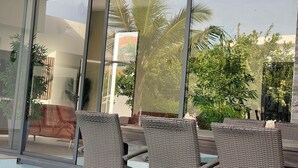 A modern outdoor seating area with wicker chairs and a glass partition, overlooking a garden with palm trees and other greenery.