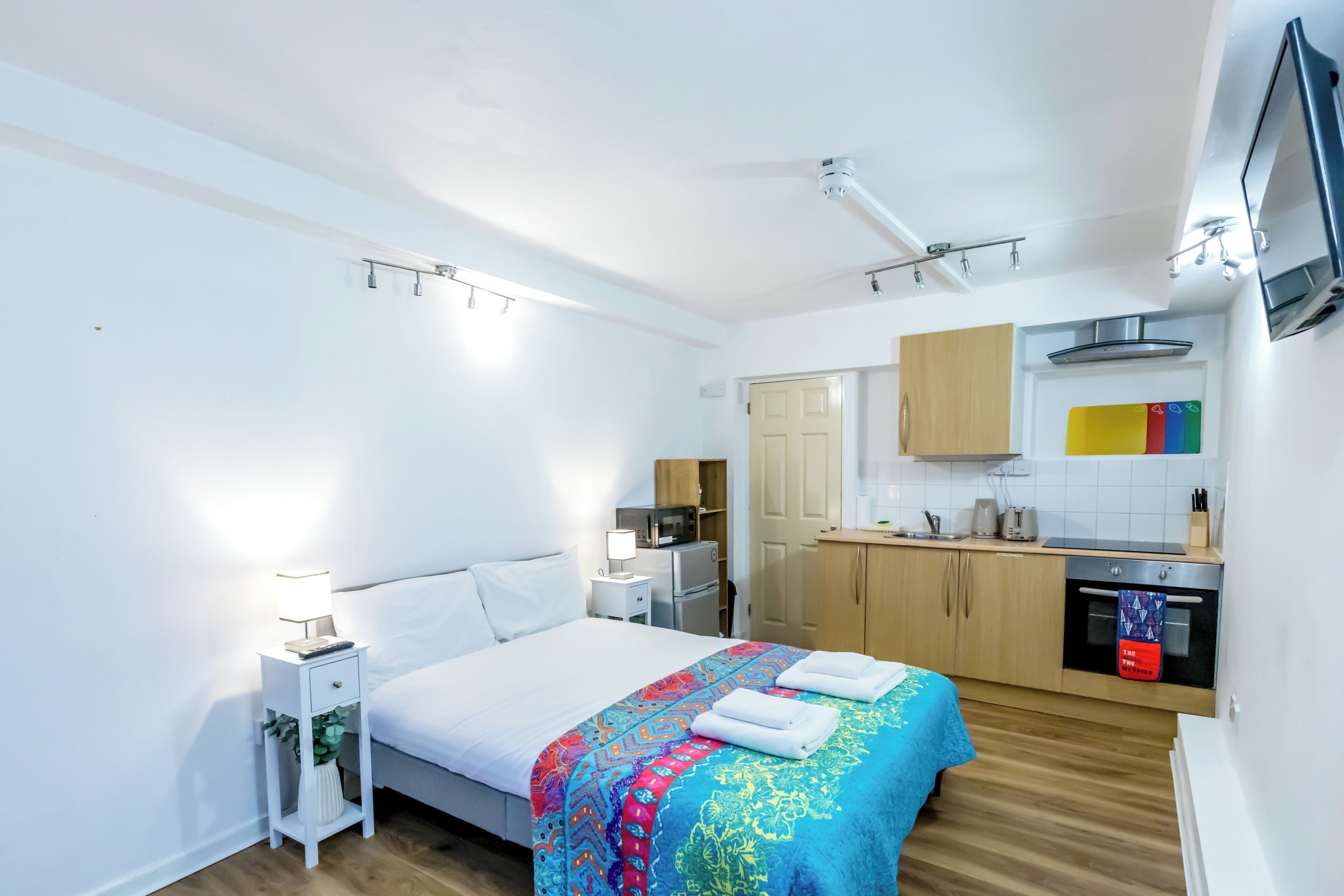 A modern hotel room with a neatly made bed, a small bedside table with a lamp, a kitchen area with wooden cabinets and a microwave, and a TV mounted on the wall.