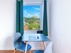 Ein Zimmer mit Tisch und zwei Stühlen, ein Fenster mit Blick auf eine hügelige Landschaft und ein kleiner Tisch mit Fernbedienung und Broschüre.