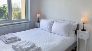 Un lit bien fait, avec du linge de lit blanc et des oreillers, deux tables de chevet avec des lampes, et une vue sur les arbres par la fenêtre.
