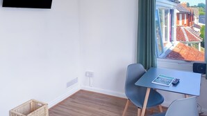 Une petite pièce avec une seule table et une seule chaise, un téléviseur fixé au mur et une fenêtre donnant sur un bâtiment au toit rouge.