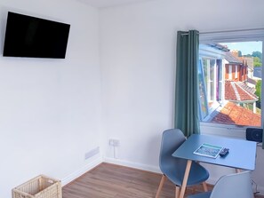 Ein kleines Zimmer mit einem Tisch, einem Stuhl, einem an der Wand befestigten Fernseher und einem Fenster mit Blick auf ein Gebäude mit rotem Dach.