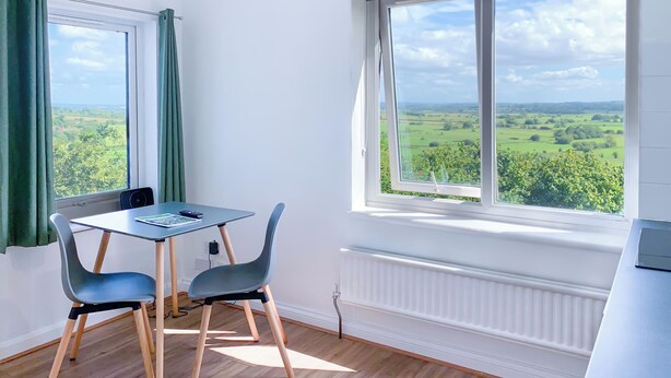 Une pièce avec une table et des chaises, un radiateur et de grandes fenêtres donnant sur des champs verdoyants.